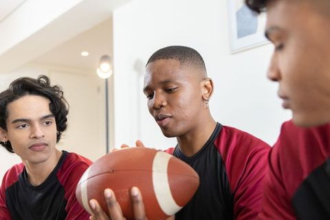 Multicultural teenage football teammates sitting indoors holding ball sharing game plan