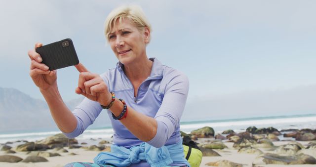 Senior woman enjoying beachfront while taking selfie with smartphone. Perfect for topics on retirement lifestyle, staying active in later years, enjoying nature, technology use among older adults, leisure activities.