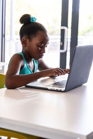 African American Girl Typing on Laptop at White Table Near Glass Doors, Studying in Daylight