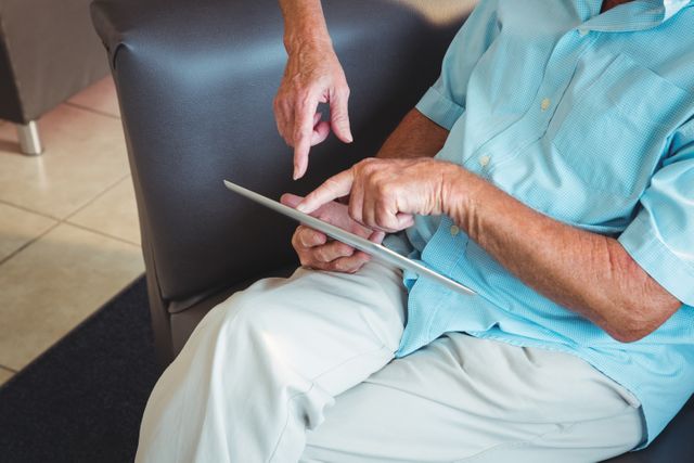 Senior man wearing casual clothes using a digital tablet with assistance from another person, likely in a retirement home. Ideal for promoting elderly tech education, senior living, modern technology adoption, healthcare support, and retirement community activities.