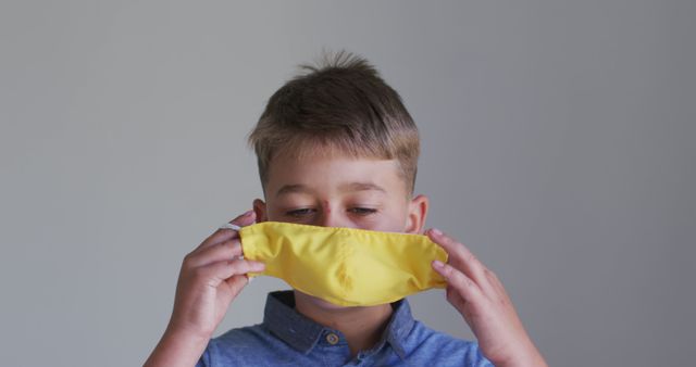 This image shows a young boy holding a yellow face mask, preparing to put it on. The child is wearing a blue shirt, and the background is plain and neutral. This image can be used for health and safety campaigns, educational materials, articles about child safety during Covid-19, or promotional content for face masks.