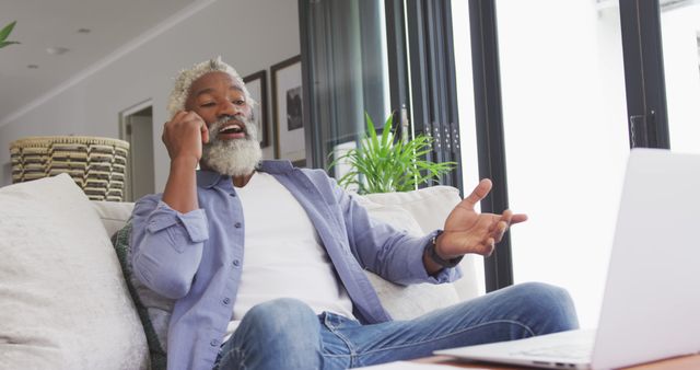 Senior man with gray hair and beard engaging in an excited conversation on his phone while sitting on a comfortable couch at home. He has a laptop in front of him, indicating he might be discussing something work-related or having a video chat. Suitable for themes like remote work, communication, home lifestyle, technology use among seniors, and positive emotions in older adults.
