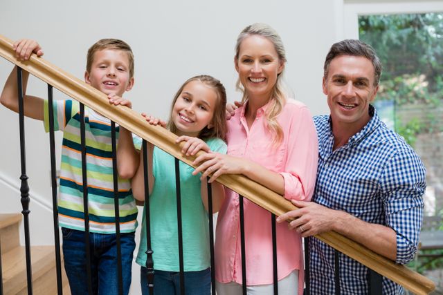 This image shows a happy family of four standing on a staircase at home, smiling and looking at the camera. It can be used for promoting family values, home living, real estate, or lifestyle content. Ideal for websites, brochures, and advertisements focusing on family life, home improvement, or residential properties.