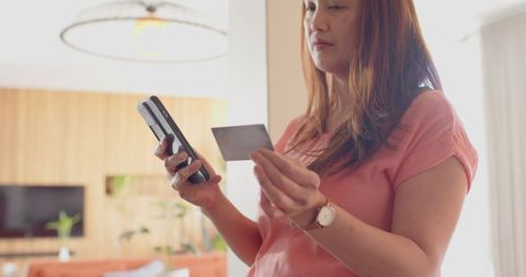 Woman in living room holding smartphone and credit card making payment
