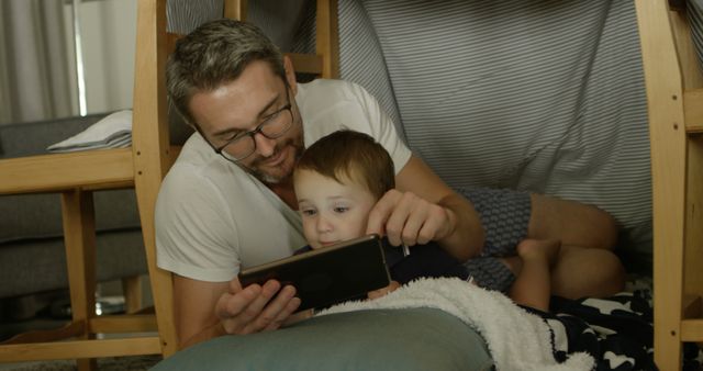 Father and young son lying inside a homemade indoor play tent and enjoying time on a tablet. The scene depicts a close family bond and casual relaxation at home. Picture perfect for use in family-oriented advertisements, parenting blogs, or technology-in-family settings depicting modern bonding experiences.
