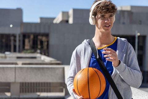 Teen male athlete holding basketball on urban rooftop wearing headphones and sports jersey