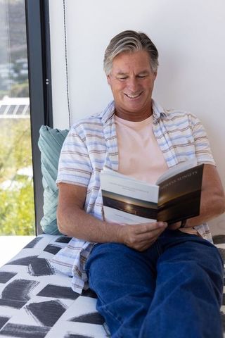 Mature man reading paperback on sunlit window seat relaxing in casual striped shirt
