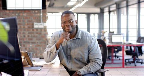 Confident professional smiling at desk in modern open-plan office with laptop and tablet