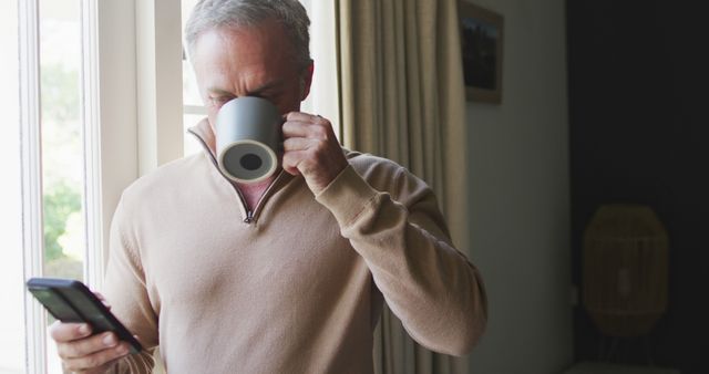 Happy caucasian man using smartphone and drinking coffee in bedroom. Spending time at home alone.