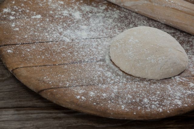 This image shows a close-up of pizza dough on a floured wooden board with a rolling pin. Ideal for use in cooking blogs, recipe websites, or advertisements for baking products. It conveys a rustic, homemade feel, perfect for illustrating the preparation process in the kitchen.