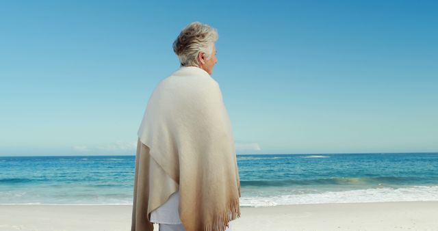 Senior woman standing on the beach with a serene expression, gazing at the ocean. She is wrapped in a shawl, symbolizing warmth and peacefulness against the clear blue sky and waves. Ideal for use in themes of tranquility, retirement, peaceful living, travel, and elderly lifestyle.