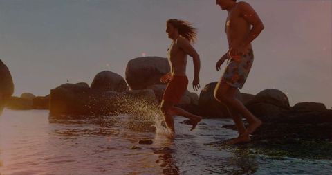 Two men running on rocky shoreline at sunset splashing water during golden hour