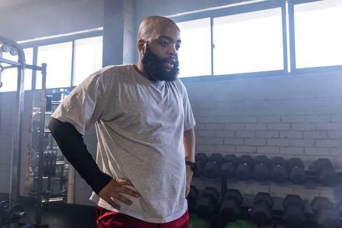 African american man catching breath after weightlifting near dumbbell rack in daylit gym