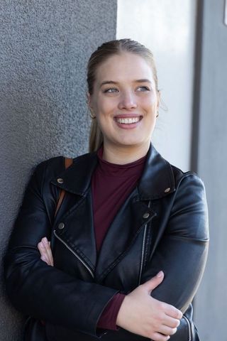 Confident young woman smiling and leaning against concrete wall in urban leather jacket