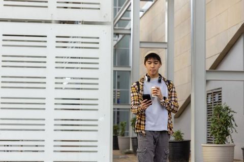 Asian man walking with coffee and smartphone in modern urban courtyard wearing beanie