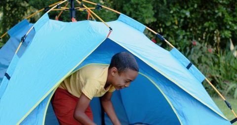 African american preteen boy popping out of bright blue pop-up tent playing in backyard