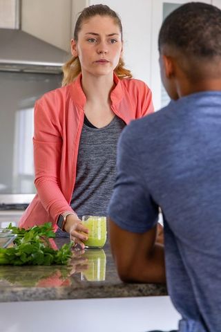 Young interracial couple standing in modern kitchen offering green smoothie on granite counter
