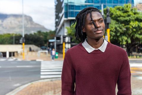Young african american man standing at urban crosswalk wearing maroon sweater, thinking