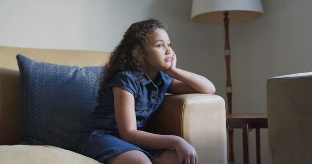 Young girl with curly hair in a denim dress sitting on a couch at home looking pensive and thoughtful. Ideal for themes of childhood, contemplation, and home life. Can be used in articles, blogs, and promotional materials focusing on family, mental health, or childhood development.
