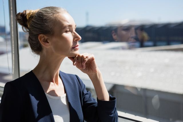 Businesswoman standing by window in office, deep in thought. Ideal for use in corporate materials, business presentations, career development guides, and articles about professional life and workplace dynamics.