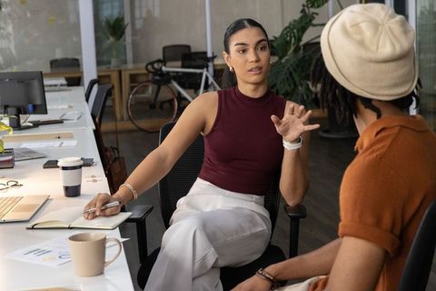 Woman leading discussion with coworker in modern open office, gesturing while taking notes