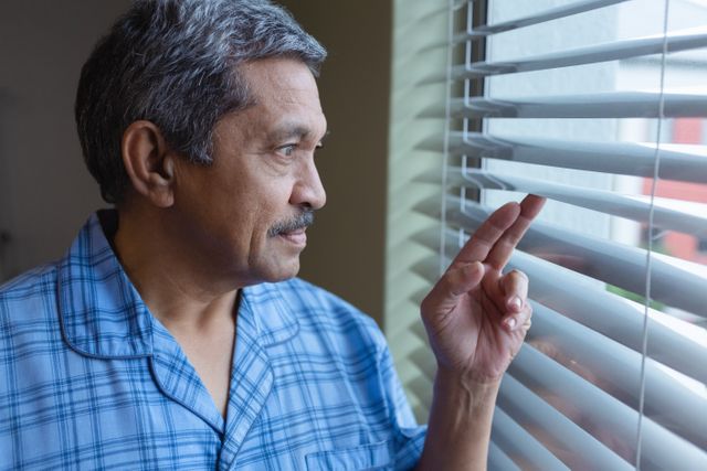 Mature male patient in hospital setting looking through window blinds, appearing thoughtful and contemplative. Ideal for use in healthcare, medical, and senior care contexts, illustrating themes of recovery, contemplation, and patient care.