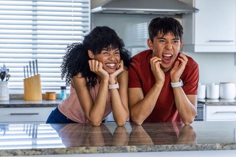 Diverse couple leaning on granite countertop reacting to smartwatch notification, joyful moment