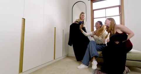 Diverse friends trying on black dresses and checking smartphone by mirror in bright wardrobe