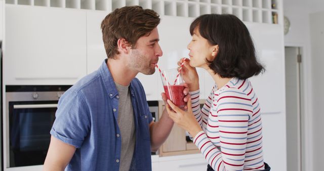 Image of happy diverse couple drinking juice with straws in kitchen. Love, relationship and spending quality time together concept.