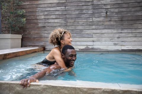 Smiling black man carrying mixed-race woman in backyard plunge pool, relaxing in modern spa