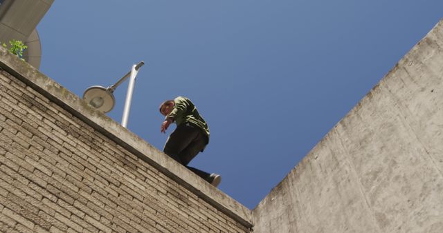 Man standing on edge of a tall building, looking down with clear blue sky in background. Suitable for themes of urban exploration, adventure, risk-taking, architectural photography, or urban lifestyle blogs. Can be used for motivational posters, brochures for safety awareness, or promotional materials for adventure sports.