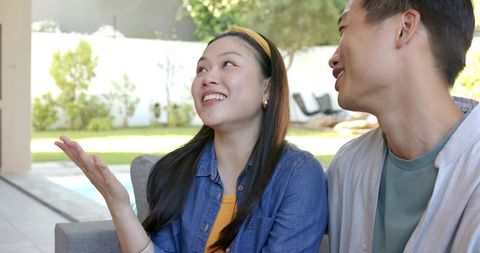 Smiling asian couple relaxing on patio couch by poolside sharing conversation and laughter
