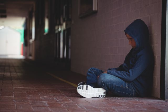 Young boy sitting alone on pavement in school corridor, looking sad and withdrawn. He is wearing a hoodie and jeans, leaning against a wall. This image can be used to depict themes of childhood loneliness, bullying, depression, and mental health issues in educational settings. Suitable for articles, blogs, and campaigns focused on child welfare, mental health awareness, and anti-bullying initiatives.
