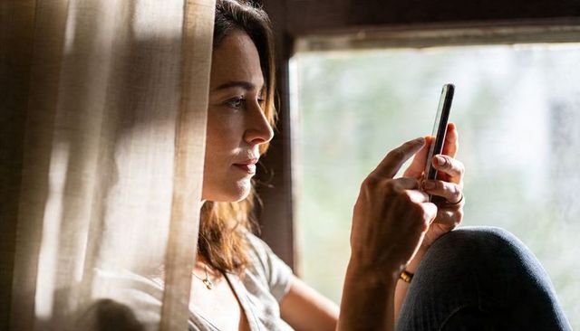 Intimate daylight portrait of woman tapping smartphone at window seat with sheer curtain