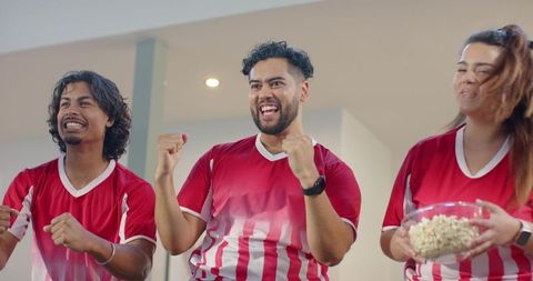 Friends cheering during live match at home wearing red and white jerseys holding popcorn