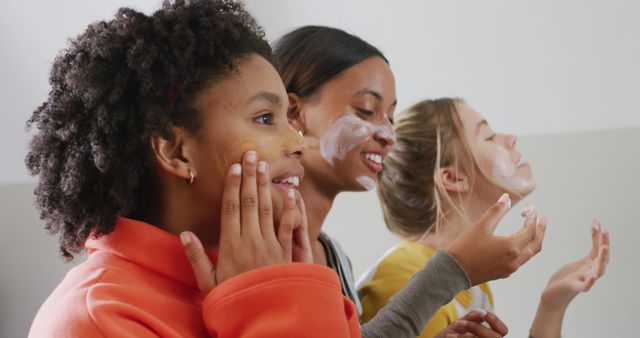 Three young women with different skin tones engaged in applying skincare products together. They are laughing and enjoying the moment, emphasizing friendship and diversity. Ideal for content related to beauty, self-care, skincare routines, and female empowerment.