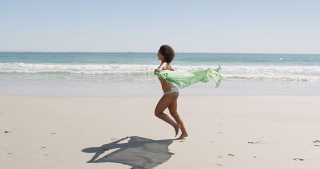 Young woman running on sandy beach, holding green scarf, creating sense of freedom and joy. Waves gently breaking in the background during clear, sunny day. Suitable for travel brochures, lifestyle blogs, advertisements for vacation destinations, or health and wellness promotions.