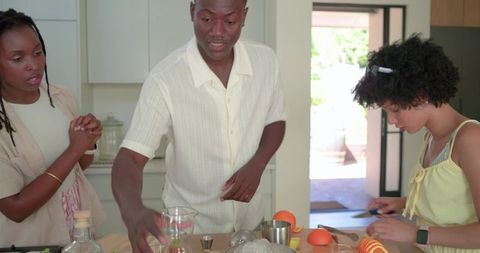 Friends mixing cocktails and slicing citrus at sunlit wooden kitchen island home gathering