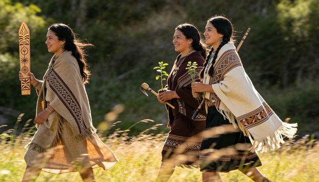 Indigenous women walking wearing woven shawls carrying ceremonial staff and carved plank