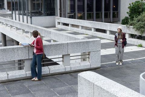 Students Leaning on Brutalist Campus Terrace Holding Coffee and Using Smartphone
