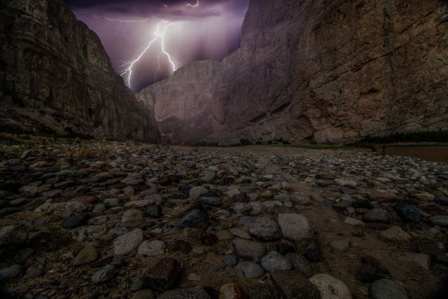 Rocky valley scene with lightning storm illuminating the night sky. Use for weather-related articles, dramatic nature visuals, or adventure themes. Emphasizes power and beauty of nature.
