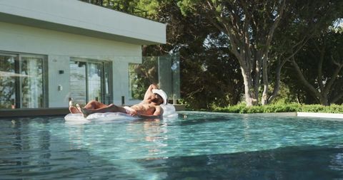 Mature man relaxing on white pool float at modern home pool, sunlit luxury lounging