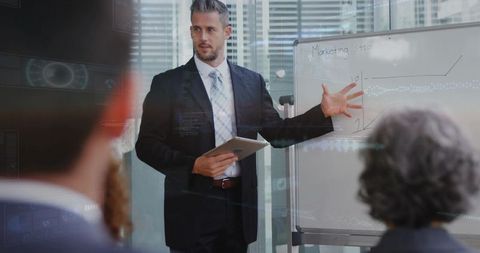 Executive presenting marketing strategy on whiteboard holding tablet in glass boardroom