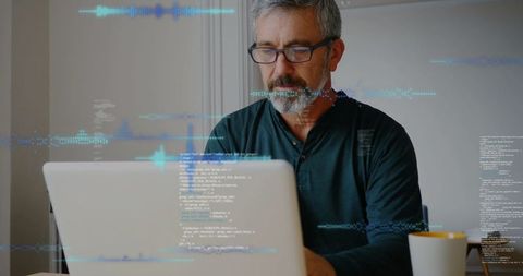 Mature professional coding on laptop in home workspace wearing green henley and eyeglasses