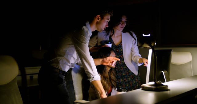 This scene shows a group of professionals collaborating in an office at night around a computer screen, emphasizing urgency and dedication in the workplace. Ideal for use in articles or resources highlighting teamwork, dedication, late-night work culture, or office environments.