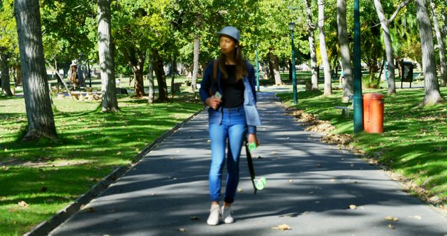 Happy biracial woman with skateboard and smartphone walking in sunny park. Free time, lifestyle, relaxation and city break, communication, unaltered.