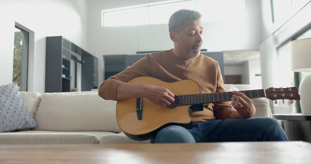 Man seated on a couch in modern living room playing acoustic guitar. Suitable for visuals related to home relaxation, casual music sessions, lifestyle, and hobbies.