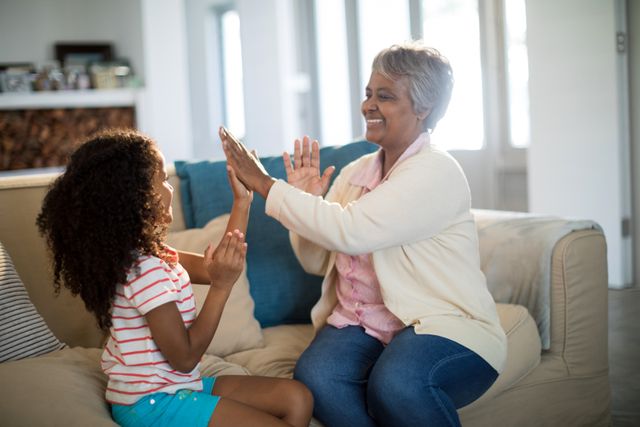 Grandmother and granddaughter enjoying a playful clapping game on a sofa in a cozy living room. Ideal for depicting family bonding, intergenerational relationships, and joyful moments at home. Perfect for use in family-oriented advertisements, articles on family life, or promotional materials for home and lifestyle products.