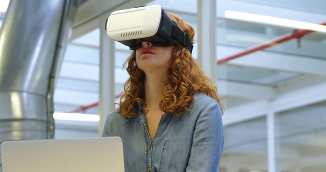 Red-haired woman using a VR headset while working at her laptop in a modern office. The scene underscores innovation and technology in a workplace. Ideal for articles or advertisements focusing on virtual reality applications in business, workplace innovation, modern office setups, or the integration of cutting-edge technology in professional environments.