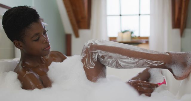 Young African American woman shaving legs while relaxing in a bubble bath at home. Capturing personal grooming and self-care. Ideal for content on beauty routines, personal hygiene, wellness, and lifestyle blogs.